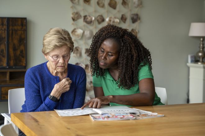 Verzorgende leest samen met oudere dame aan tafel een tijdschrift