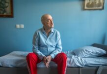 man met rode broek aan en blauw overhemd zit op bed en kijkt naar buiten